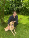Man with glasses and a dog sitting on grass with trees in the background